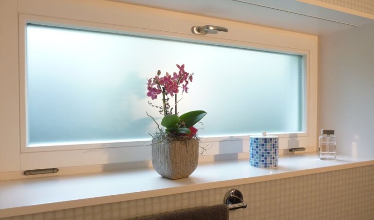 Innenaufnahme eines modernen Badezimmers mit grossflächigem Fenster und milchiger Sichtschutzfolie. Die gleichmässig matte Folie verhindert unerwünschte Einblicke von aussen und lässt gleichzeitig viel natürliches Tageslicht in den Raum. Der weisse Fensterrahmen, die dekorative Orchidee und die hellen Fliesen unterstreichen die freundliche und elegante Atmosphäre.