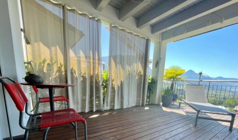 Aussenaufnahme einer überdachten Terrasse mit hellen, halbtransparenten Balkonvorhängen. Die bodenlangen Stoffbahnen sind an einer deckenmontierten Vorhangschiene befestigt und bieten flexiblen Sicht- und Sonnenschutz, ohne die Terrasse vollständig abzudunkeln. Die gemütliche Sitzgruppe mit roten Stühlen, die Sonnenliege und der beeindruckende Blick auf die See- und Berglandschaft schaffen eine einladende Atmosphäre im Aussenbereich.
