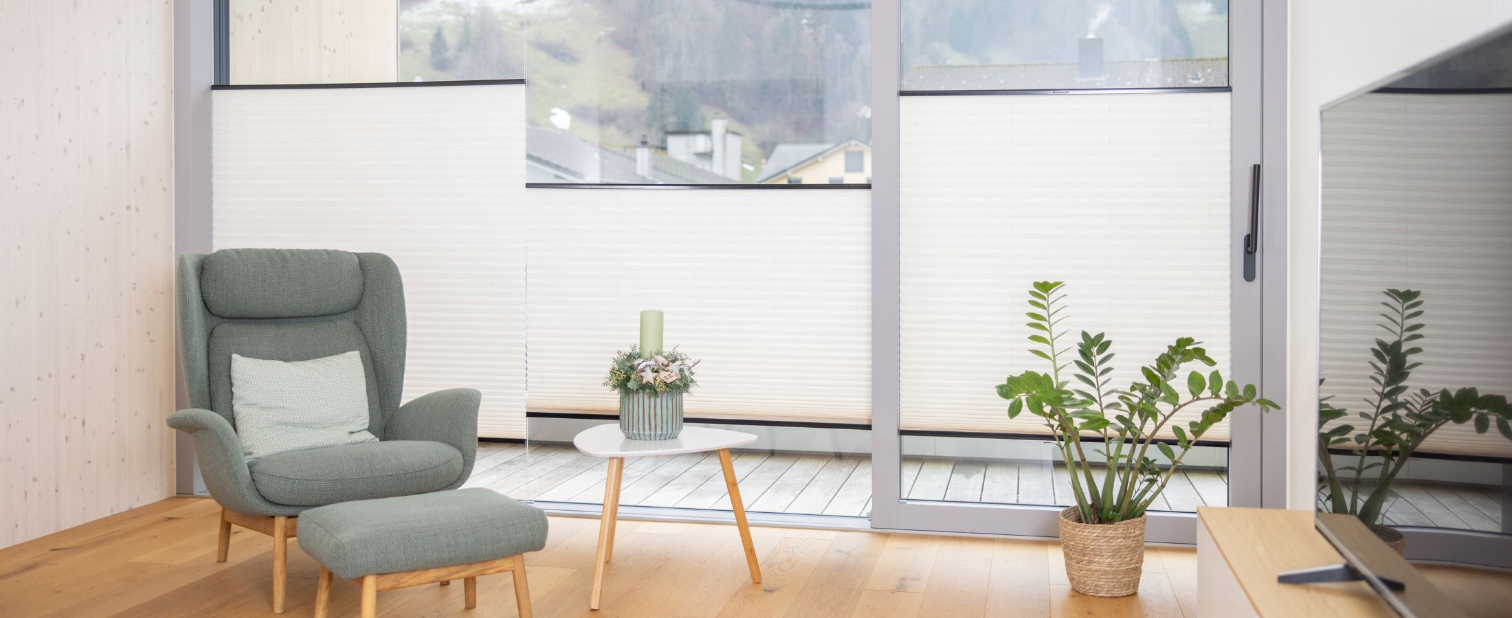 Heller Wohnraum mit grossflächiger Fensterfront und weissen Plissees als Sichtschutz im unteren Fensterbereich. Ein grüner Sessel mit Hocker, ein kleiner Beistelltisch und Zimmerpflanzen sorgen für eine gemütliche Atmosphäre